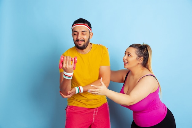 Joven pareja bastante caucásica en la formación de ropa brillante sobre fondo azul Concepto de deporte, emociones humanas, expresión, estilo de vida saludable, relación, familia. Entrenando con pesas, diviértete.