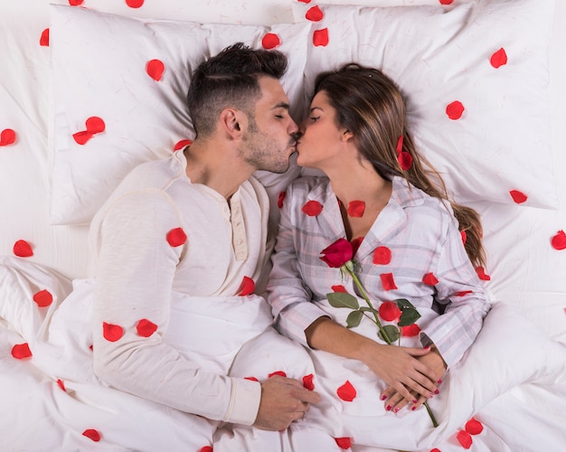 Joven pareja besándose en la cama con pétalos de rosa