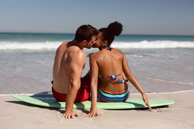 Joven pareja besándose mientras está sentado en la tabla de surf en la playa bajo el sol