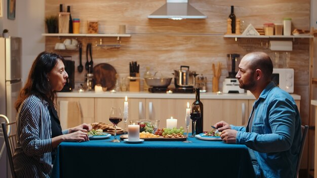 Joven pareja casada feliz con videollamada en la cocina durante la cena romántica, comiendo comida festiva. Punto de vista en línea Internet conferencia moderna, chat, comunicación, conversación de chat llamada a través de cámara web