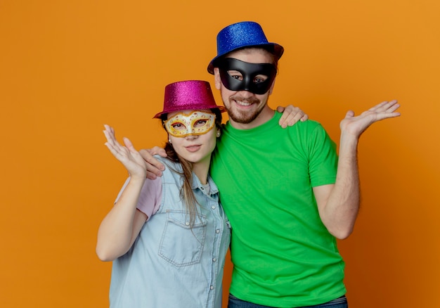 Foto gratuita joven pareja complacida con sombreros rosados y azules se puso máscaras de ojos de mascarada levantando la mano mirando aislado en la pared naranja