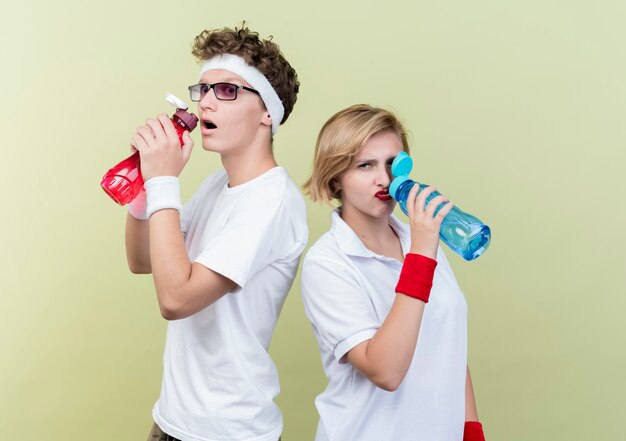 Joven pareja deportiva hombre y mujer bebiendo agua después del entrenamiento de pie sobre una pared de luz