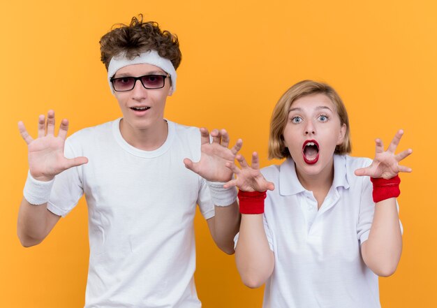 Joven pareja deportiva hombre y mujer con manos sonriendo divirtiéndose de pie sobre la pared naranja