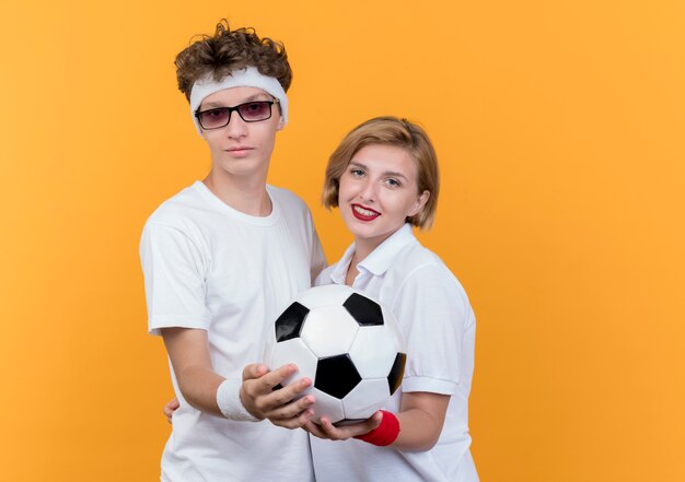 Joven pareja deportiva hombre y mujer de pie juntos sosteniendo un balón de fútbol sonriendo sobre la pared naranja