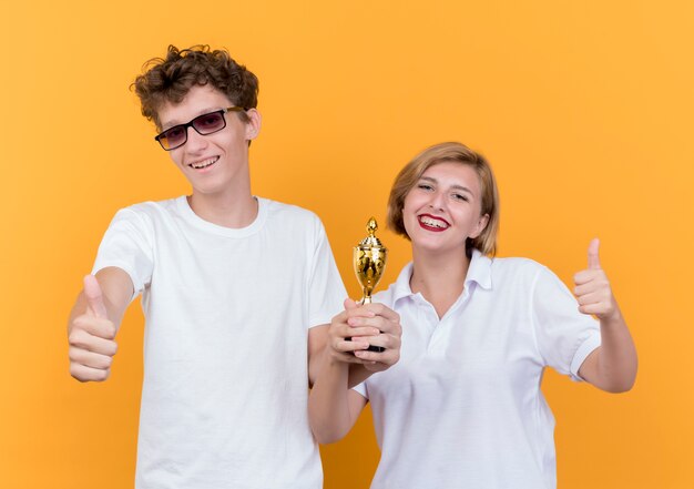 Joven pareja deportiva hombre y mujer de pie juntos sosteniendo el trofeo mostrando los pulgares para arriba sonriendo de pie sobre la pared naranja