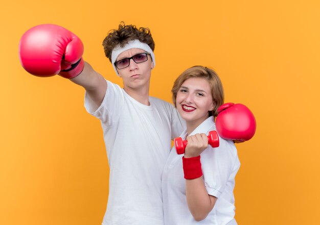 Joven pareja deportiva mujer con pesas y hombre con guantes de boxeo sonriendo de pie sobre la pared naranja