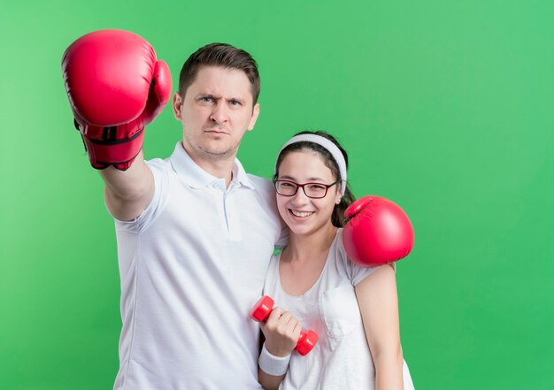 Joven pareja deportiva mujer con pesas y hombre con guantes de boxeo sonriendo de pie sobre la pared verde