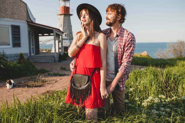 Joven pareja elegante enamorada en el campo, estilo bohemio indie hipster, vacaciones de fin de semana, traje de verano, vestido rojo, hierba verde, tomados de la mano, sonriendo