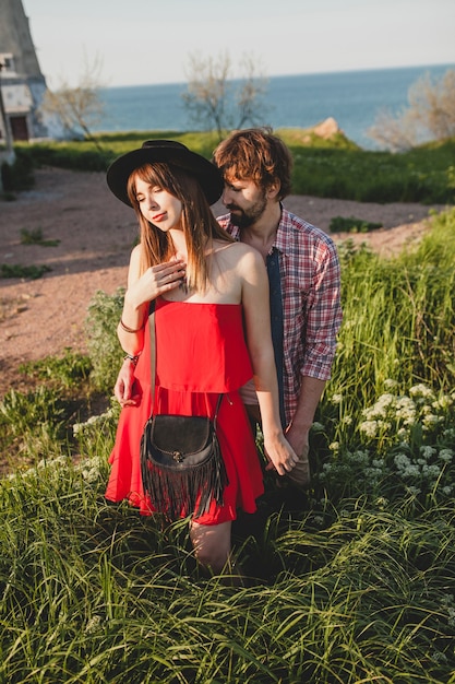 Joven pareja elegante enamorada en el campo, estilo bohemio indie hipster, vacaciones de fin de semana, traje de verano, vestido rojo, hierba verde, tomados de la mano, sonriendo