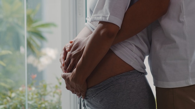 Joven pareja embarazada de Asia abraza y sosteniendo el vientre hablando con su hijo. Mamá y papá se sienten felices sonriendo pacíficos mientras cuidan bebé, embarazo cerca de la ventana en la sala de estar en casa.