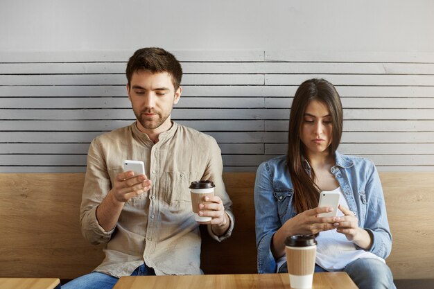 Joven pareja de estudiantes sentados en una cita en comida rápida, tomando café, mirando en teléfonos celulares, ignorándose mutuamente después de una discusión