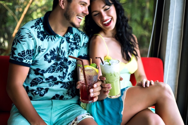 Joven pareja feliz bebiendo sabrosos cócteles dulces en el bar tropical, sonriendo y divirtiéndose, ropa brillante y emociones positivas.