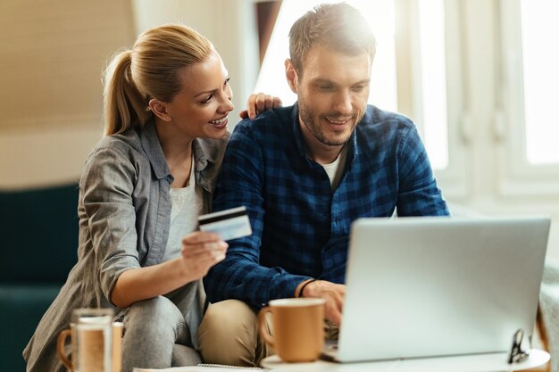 Joven pareja feliz comprando en casa mientras usa computadora y tarjeta de crédito.