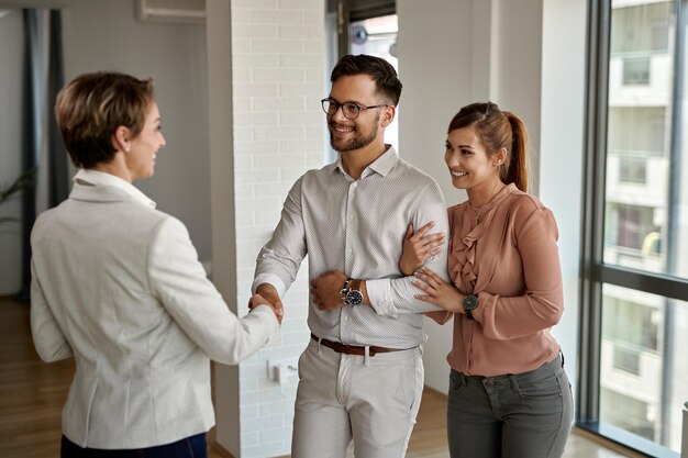 Una joven pareja feliz dándose la mano con un agente inmobiliario mientras compra un apartamento nuevo