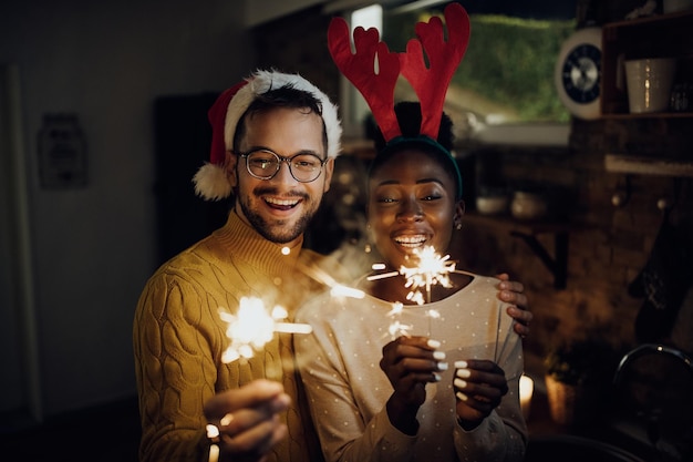 Joven pareja feliz divirtiéndose con bengalas en la víspera de Navidad en casa