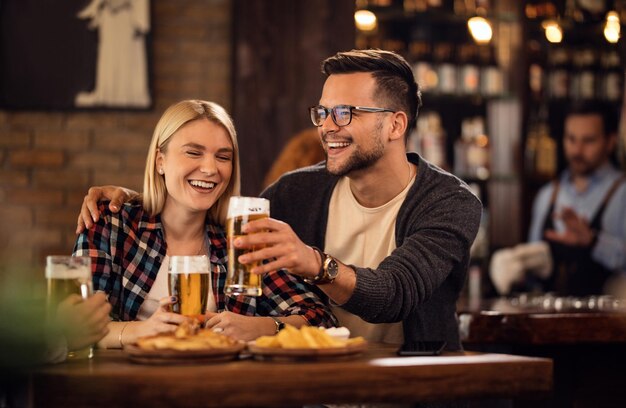 Una joven pareja feliz divirtiéndose juntos mientras brindan con cerveza en un bar