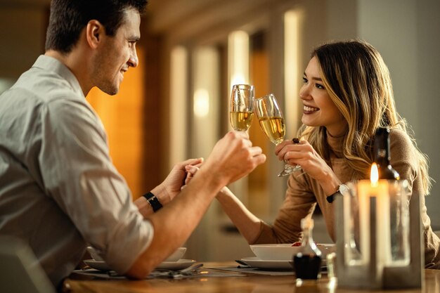 Joven pareja feliz enamorada brindando con champán mientras se toma de la mano en la mesa del comedor El foco está en la mujer