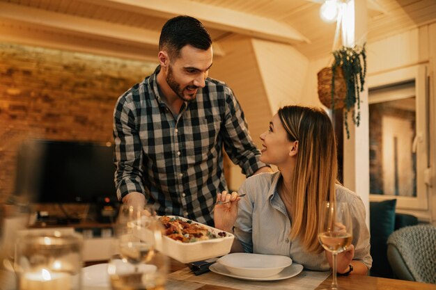 Joven pareja feliz hablando mientras el hombre sirve comida en la mesa del comedor