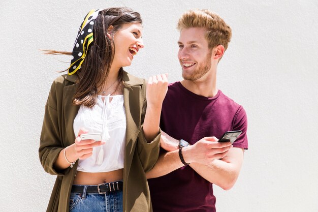 Joven pareja feliz mirando el uno al otro sosteniendo el teléfono celular contra el fondo blanco