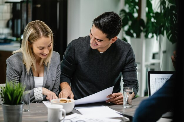 Joven pareja feliz pasando por su proyecto de vivienda en un plano durante la reunión con el agente inmobiliario