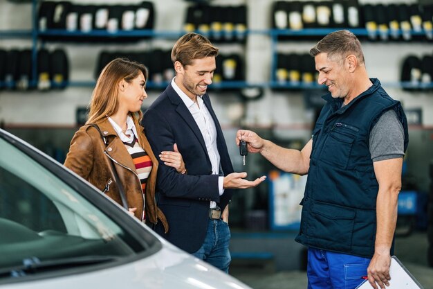 Joven pareja feliz recibiendo la llave del auto de un mecánico en un taller de reparación de automóviles