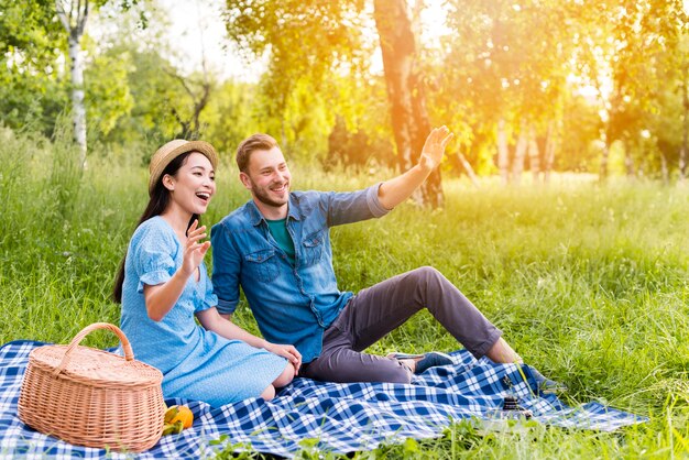 Joven pareja feliz saludando y sonriendo en picnic en la naturaleza