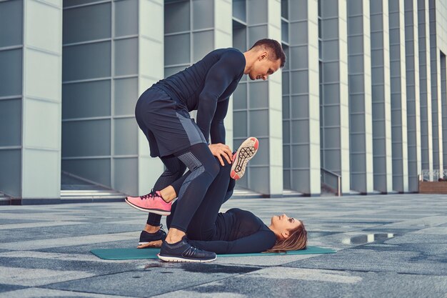 Joven pareja de fitness en ropa deportiva, haciendo estiramientos mientras se prepara para un ejercicio serio en la ciudad moderna contra un rascacielos.