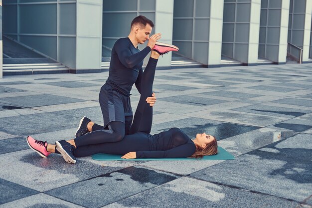 Joven pareja de fitness en ropa deportiva, haciendo estiramientos mientras se prepara para un ejercicio serio en la ciudad moderna contra un rascacielos.