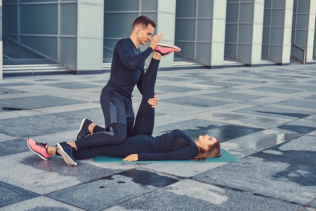 Joven pareja de fitness en ropa deportiva, haciendo estiramientos mientras se prepara para un ejercicio serio en la ciudad moderna contra un rascacielos.