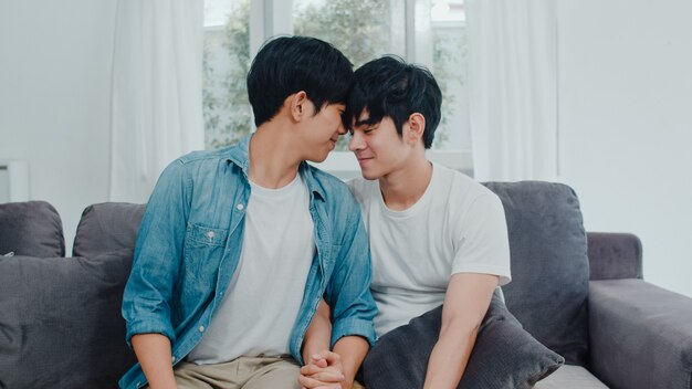 Joven pareja gay asiática abrazo y beso en casa. Los hombres de orgullo LGBTQ asiáticos y felices se relajan felices y pasan un momento romántico juntos mientras descansan en el sofá de la sala.