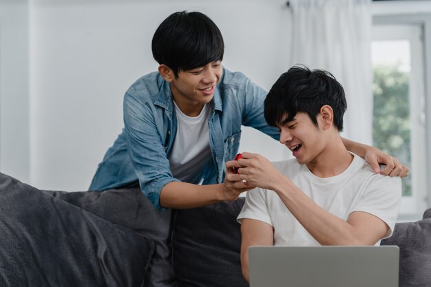Joven pareja gay asiática propone en casa moderna, los hombres LGBTQ coreanos adolescentes sonrientes felices tienen un momento romántico mientras proponen y el matrimonio sorpresa usa el anillo de bodas en la sala de estar de la casa.