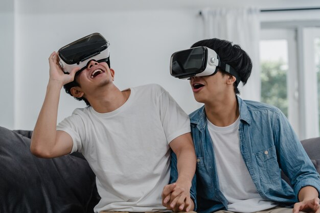 Joven pareja gay asiática que usa tecnología divertida en casa, un chico amante de Asia lgbtq + se siente feliz y divertido en realidad virtual, VR jugando juntos mientras está acostado en el sofá de la sala de estar en casa.