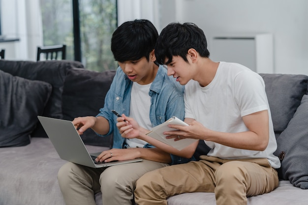 Joven pareja gay asiática trabajando portátil en casa moderna. Asia LGBTQ + hombres felices relajarse divertirse usando la computadora y analizar sus finanzas en internet juntos mientras se está acostado en el sofá de la sala de estar en casa.