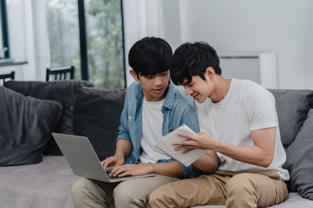 Joven pareja gay asiática trabajando portátil en casa moderna. Asia LGBTQ + hombres felices relajarse divertirse usando la computadora y analizar sus finanzas en internet juntos mientras se está acostado en el sofá de la sala de estar en casa.