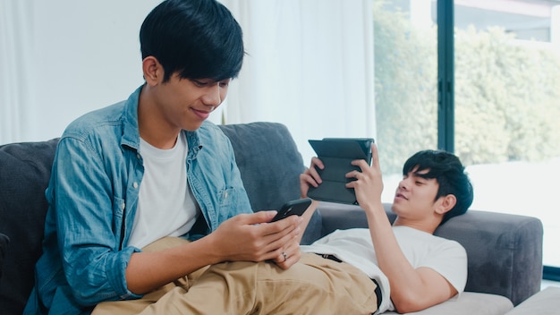 Joven pareja gay lgbtq utilizando teléfono móvil y tableta en casa moderna. Hombre asiático amante feliz relajarse risa y diversión tecnología jugar juegos en internet juntos mientras está acostado sofá en la sala de estar.