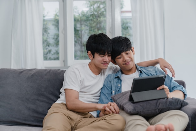 Joven pareja gay con tableta en casa. Los hombres asiáticos LGBTQ + felices se divierten y se divierten usando la tecnología para ver películas en Internet juntos mientras descansan en el sofá de la sala.