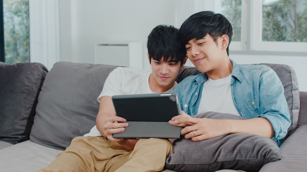 Joven pareja gay con tableta en casa. Los hombres asiáticos LGBTQ + felices se divierten y se divierten usando la tecnología para ver películas en Internet juntos mientras descansan en el sofá de la sala.