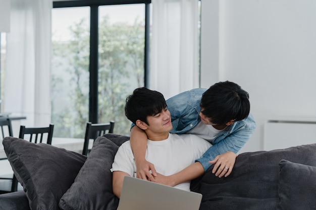 Joven pareja gay usando la computadora portátil en casa moderna. los hombres asiáticos lgbtq + felices se divierten y se divierten usando la tecnología para ver películas en internet juntos mientras descansan en el sofá de la sala de estar de la casa.