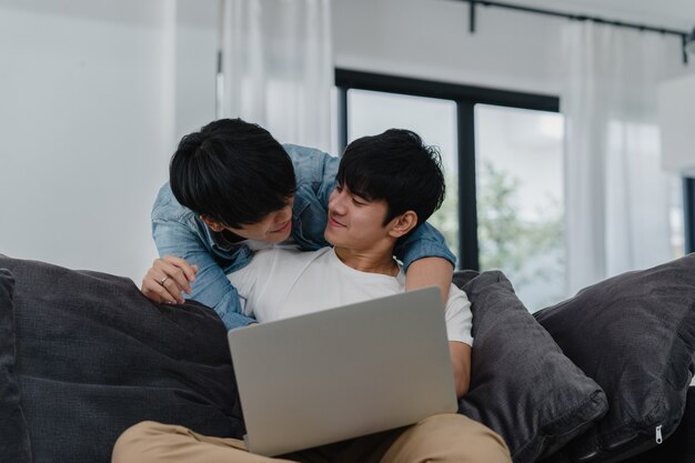 Joven pareja gay usando la computadora portátil en casa moderna. Los hombres asiáticos LGBTQ + felices se divierten y se divierten usando la tecnología para ver películas en Internet juntos mientras descansan en el sofá de la sala de estar de la casa.