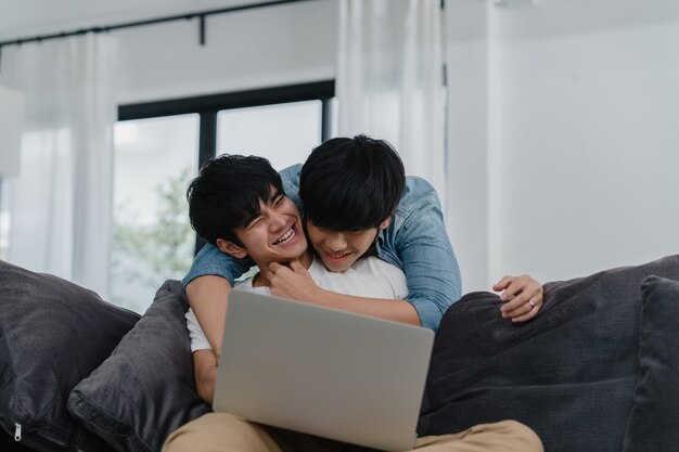 Joven pareja gay usando la computadora portátil en casa moderna. Los hombres asiáticos LGBTQ + felices se divierten y se divierten usando la tecnología para ver películas en Internet juntos mientras descansan en el sofá de la sala de estar de la casa.