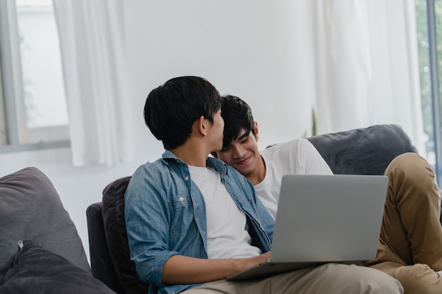 Joven pareja gay usando la computadora portátil en casa moderna. Los hombres asiáticos LGBTQ + felices se divierten y se divierten usando la tecnología para ver películas en Internet juntos mientras descansan en el sofá de la sala de estar de la casa.