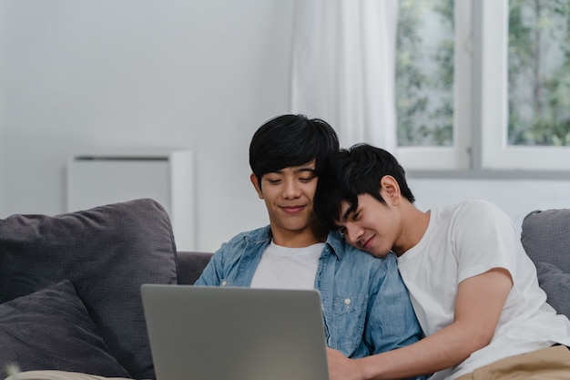 Joven pareja gay usando la computadora portátil en casa moderna. Los hombres asiáticos LGBTQ + felices se divierten y se divierten usando la tecnología para ver películas en Internet juntos mientras descansan en el sofá de la sala de estar de la casa.