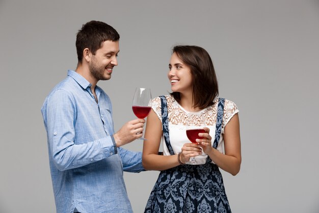 Joven pareja hermosa bebiendo vino, sonriendo sobre la pared gris