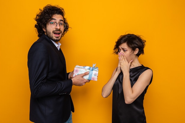 Joven pareja hermosa feliz dando un regalo a su novia sorprendida y asombrada celebrando el día internacional de la mujer el 8 de marzo de pie sobre fondo naranja