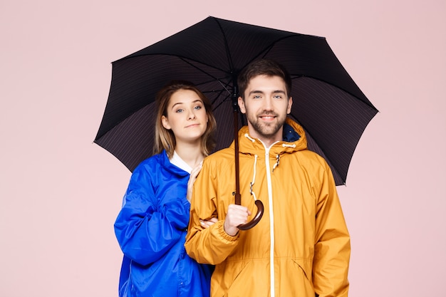 Joven pareja hermosa posando en abrigos de lluvia con paraguas sobre pared rosa claro