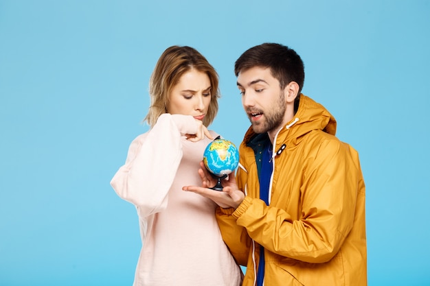 Joven pareja hermosa posando apuntando a diferentes lugares del pequeño globo sobre la pared azul Hombre con abrigo de lluvia.