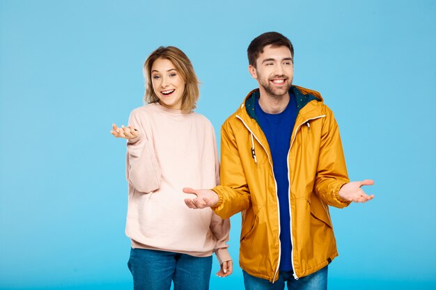 Joven pareja hermosa posando sonriendo divirtiéndose sobre pared azul Hombre con abrigo de lluvia.