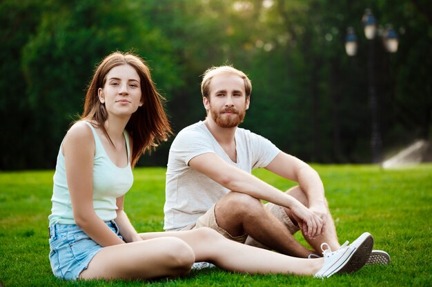 Joven pareja hermosa sonriendo, sentado en el césped en el parque.