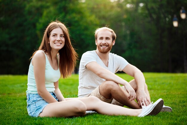Joven pareja hermosa sonriendo, sentado en el césped en el parque.