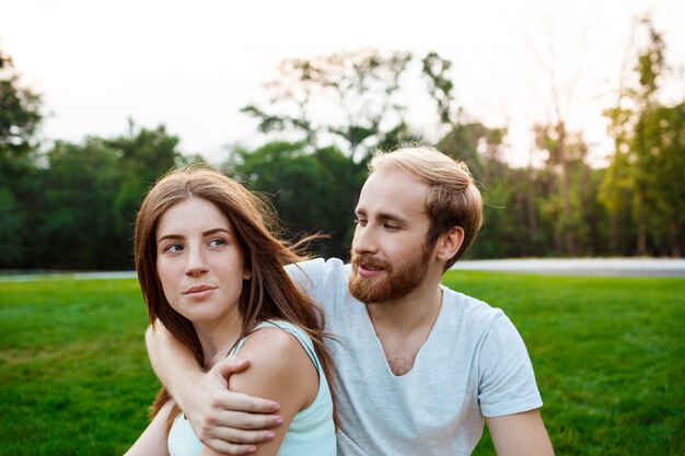 Joven pareja hermosa sonriendo, sentado en el césped en el parque.